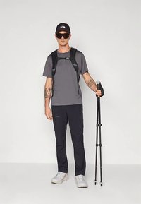 Pelēka īsroku krekls, melnas bikses un baltas sporta kurpes. Valkā melnu cepuri un saulesbrilles; turot pārgājienu nūjas. Minimālistisks dizains.