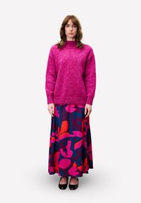 Femme avec des cheveux bruns ondulés portant un pull rose vif et une longue jupe sombre avec des motifs floraux abstraits roses et rouges, se tenant droite.