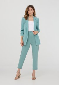 Blazer verde acqua e pantaloni sartoriali abbinati in un tessuto morbido. I pantaloni sono dotati di chiusura frontale con bottone. Il modello indossa una canotta bianca e scarpe con tacco a strappo.