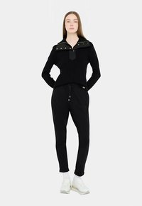 Must soonikuga kampsun kontrastse krae ja nööpdetailidega, koos mustade jookspükstega. Modell kannab valgeid tenniseid sokkidega.