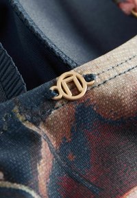 Stoffdetail mit einem gemusterten Design in Dunkelblau-, Rot- und Brauntönen, mit einem goldfarbenen Metallakzent-Knopf.