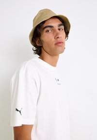Hvid bomulds t-shirt med korte ærmer, der har et BMW-logo og tekst. Kombineret med en lysbrun bucket hat og styling med bølget hår.