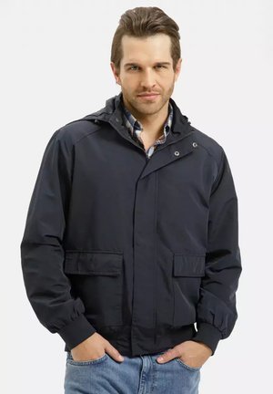 Mężczyzna w ciemnogranatowej kurtce na koszulę w kratę, z rękami w kieszeniach, stoi na jasnym, jednolitym tle.