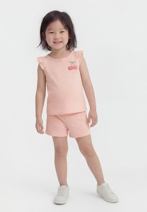 Mladé dievča stojí a usmieva sa, má na sebe svetloružové tielko bez rukávov s motívom čerešní a zodpovedajúce šortky, biele tenisky na bielom pozadí.