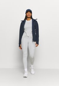 Conjunto de vestuário ativo com uma jaqueta navy com fecho, uma t-shirt cinza com nó, leggings cinza claro e ténis brancos com detalhes canelados.