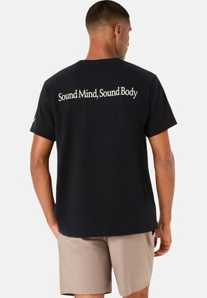 Mann trägt ein schwarzes T-Shirt mit dem Schriftzug „Sound Mind, Sound Body“ auf dem Rücken und beigen Shorts, steht mit dem Rücken zum Betrachter auf weißem Hintergrund.