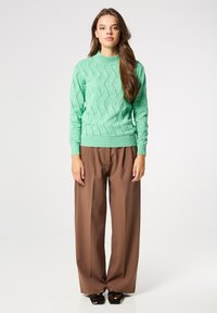 Suéter de punto verde menta con un patrón de olas texturizado, cuello alto y puños acanalados, combinado con pantalones anchos marrones y calzado oscuro.