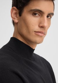 Maglione nero a collo alto in maglia con texture a coste e dettagli delle cuciture sottili. Veduta ravvicinata che mette in risalto il collo e la qualità del tessuto.