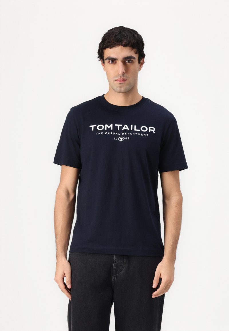 Tamsiai mėlynas medvilninis marškinėliai trumpomis rankovėmis ir apvaliu iškirpimu, su baltais spausdintais žodžiais: "TOM TAILOR THE CASUAL DEPARTMENT 1962."