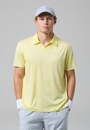 Giovane uomo che indossa una polo giallo chiaro, un cappellino grigio chiaro, polsini bianchi e pantaloncini grigio chiaro, in piedi con le mani nelle tasche.