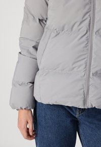 Grå vadderad jacka med dragkedja fram, rundad krage och detaljer med elastiska ärmslut. Matchad med blå denimjeans för kontrast.