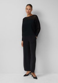 Maglione nero a coste con maniche trasparenti a righe, abbinato a pantaloni neri larghi e décolleté nere con punta appuntita. Texture liscia, design aderente.