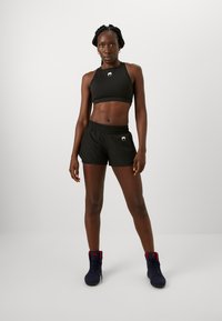 Top cropped atlético preto e calções com cintura elástica, apresentando um logótipo. Pessoa usa ténis azuis com detalhes vermelhos. Textura suave.