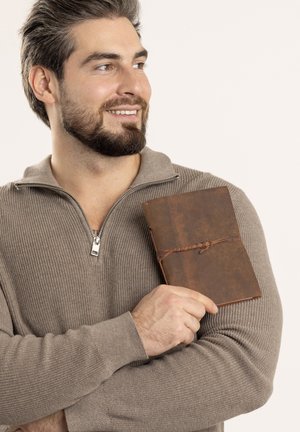 Ledergebundenes Notizbuch mit einer strukturierten braunen Einband, gesichert mit einer Schnurwicklung, gehalten vor einer Person, die einen hellgrauen Strickpullover trägt.