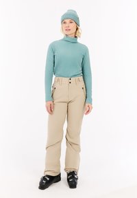 Col roulé côtelé bleu clair, pantalons de ski beiges avec deux poches avant et quincaillerie noire, accompagnés d'un bonnet tricoté assorti et de bottes de ski noires.