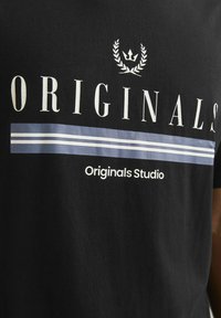 Camiseta de algodón negra con el texto en blanco "ORIGINALS" y "Originals Studio" en negrita, con una franja horizontal azul. Textura suave, corte estándar.