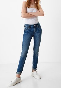 Slim-fit blue jeans with a faded finish, five-pocket design, and rolled cuffs, paired with white sneakers and a fitted white tank top.