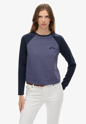 Joven mujer con cabello largo castaño que lleva una camiseta de manga larga azul y marino con el texto "SUPERDRY", pantalones blancos y un cinturón trenzado marrón.