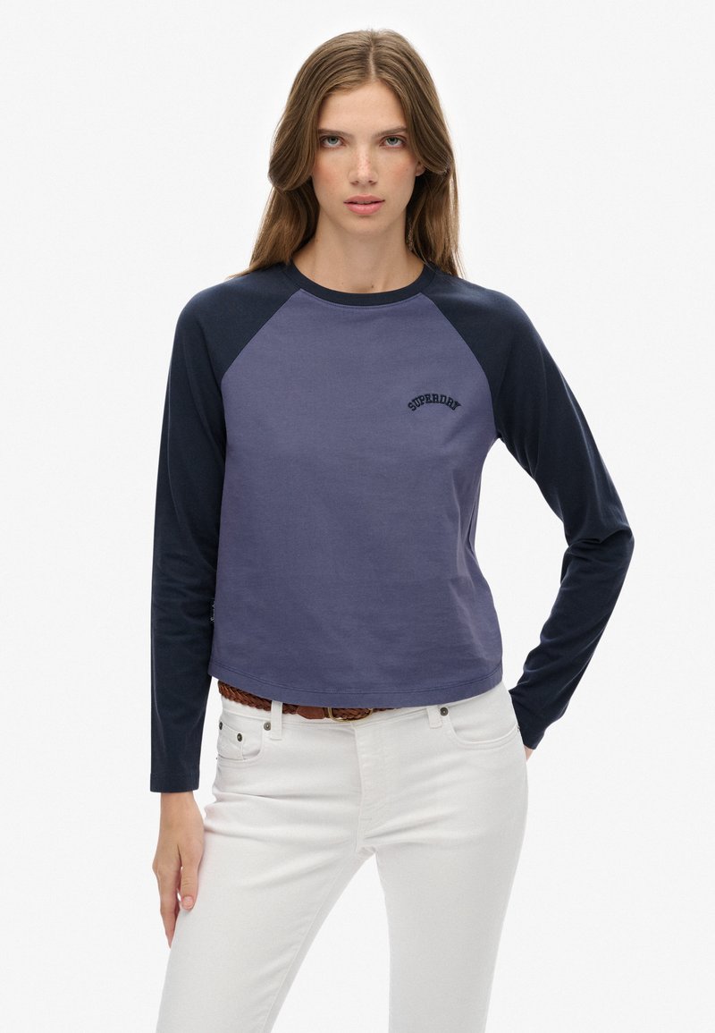 Jonge vrouw met lang bruin haar, kleed in een blauw en marineblauw shirt met lange mouwen met de tekst "SUPERDRY", witte broek en een leren gevlochten riem.