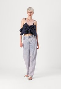 Mornarsko modra majica s kratkimi rokavi z velikim pentljo, v kombinaciji s svetlimi, ohlapnimi kavbojkami. Model nosi ravne sandale, kar poudarja minimalističen stil.