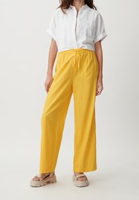 Geel wijde broek met een elastische tailleband en trekkoord, gecombineerd met een witte overhemd met kraag en beige platform sandalen.