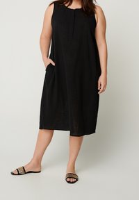 Černé bezrukávové šaty s volným střihem, textura materiálu a boční kapsy. Kombinováno s černými klouzavými sandály s tkaným páskem.