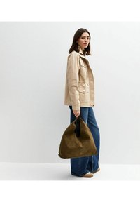 Beige canvas jack met zakken, gecombineerd met wijde blauwe jeans. Model houdt een suède olijfgroen tasje vast. Eenvoudig, casual ontwerp.