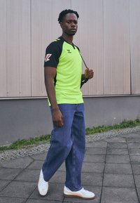 Maglietta sportiva verde neon con maniche nere e dettagli gialli, abbinata a jeans azzurri larghi e sneakers bianche con dettagli color beige.