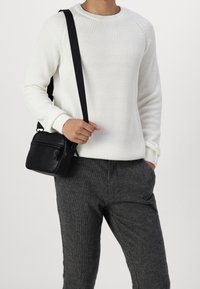 Black crossbody bag with zipper, smooth texture, worn with a white ribbed sweater and gray patterned trousers. Simple, functional design.