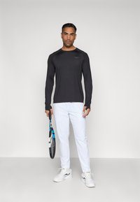 Ilgomis rankovėmis juoda sportinė marškinė su apvaliu iškirpimu, derinamos su baltomis kelnėmis ir baltais sportbačiais; laiko teniso raketę.