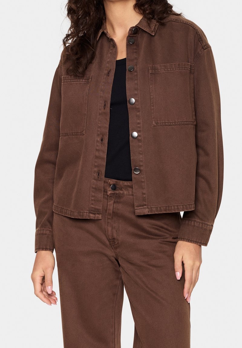 Veste en denim marron avec col, boutons sur le devant et deux poches poitrine. Portée sur un haut noir, elle présente une coupe décontractée et une texture lisse.