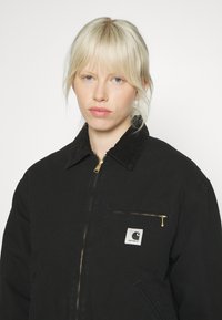 Schwarze Stoffjacke mit Reißverschluss und Cordkragen. Verfügt über eine Fronttasche mit goldfarbenem Reißverschluss und Markenetikett im linken Brustbereich.