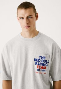 Graues Baumwoll-T-Shirt mit Rundhalsausschnitt, ausgestattet mit einer Brusttasche, die den blauen und roten Text "THE RED BULL RACING TEAM EST. 2004" trägt.