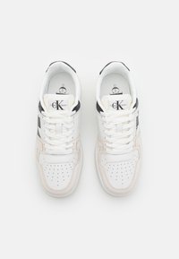 Ett par vita låga sneakers med beige mockadetaljer, svart häl, vita snören och Calvin Klein Jeans-logotyp på tungan och innersulan.