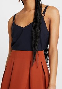 Tmavě modrý top na ramínka s výstřihem do V a tenkýmiramínky, s bronzovými doplňky; v kombinaci s sukní s plisováním v barvě rezavé.
