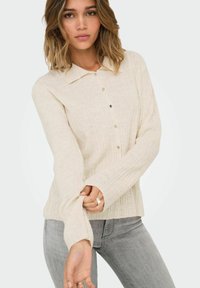Cremefarbiges, geripptes Shirt mit Kragen, Knopfleiste und langen Ärmeln; weiche Textur mit subtilen vertikalen Linien. Kombiniert mit grauen Jeans.