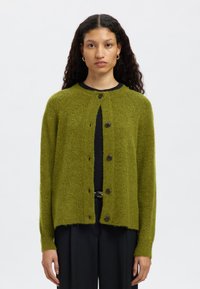 Žalias, tekstūruotas kardiganas su apvalia iškirpte, sagomis priekyje ir ilgomis rankovėmis, derinamas su juoda palaidine ir tamsiomis kelnėmis.