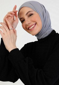 Hijab grigio, maglia nera scintillante a maniche lunghe. Mani adornate con anelli d'argento. Sfondo chiaro e neutro. Espressione sorridente.