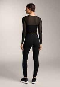 Fekete, testre simuló, hosszú ujjú crop top áttetsző felső résszel, magas derekú fekete leggingssel és fekete sportcipővel párosítva. Sima textúra, minimalista kialakítás.