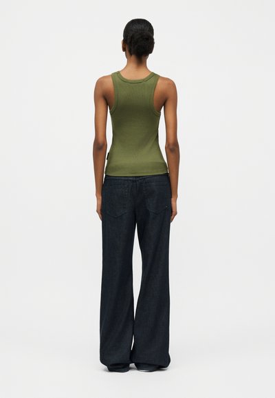 Woman with dark skin and hair in bun wearing green sleeveless top and loose dark blue jeans, standing facing away against white background.