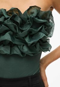 Tmavě zelený body s velkými, vrstvenými volány u výstřihu. Tkanina má hladkou strukturu s jemným leskem a žebrovaným vzorem dole.