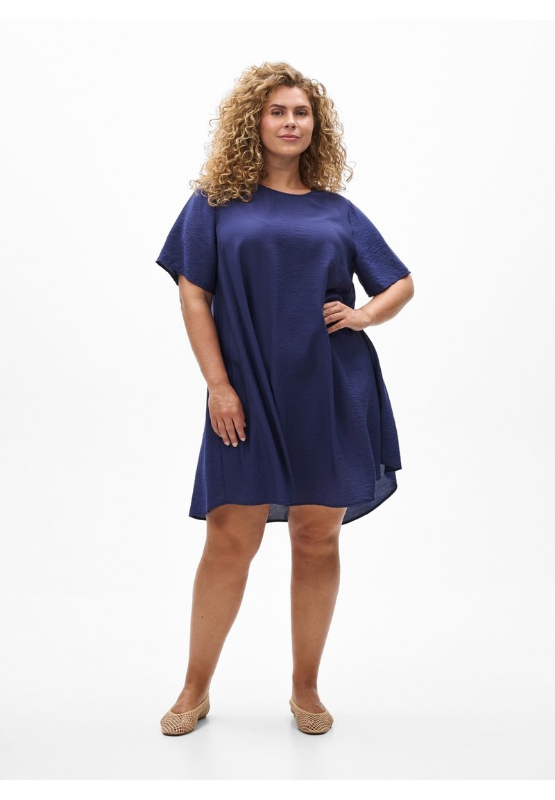Mujer con cabello rizado, vestida con un vestido suelto azul marino y zapatos planos beige, de pie con una mano en la cadera contra un fondo blanco.