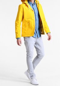 Giacca impermeabile gialla con zip frontale, cappuccio e tasche con bottone, indossata sopra una camicia azzurro chiaro, abbinata a pantaloni grigio chiaro e sneakers bianche.