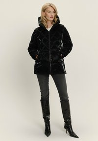 Zwarte pufferjack met een glanzende afwerking, ruitpatroon, capuchon en ritssluiting aan de voorkant; gecombineerd met zwarte skinny jeans en laarzen tot de knie.