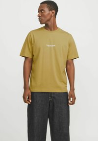 Olive green cotton t-shirt with short sleeves, featuring the “Originals Studio” logo in white on the chest. Paired with dark denim trousers.