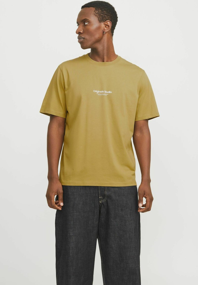 Olive green cotton t-shirt with short sleeves, featuring the “Originals Studio” logo in white on the chest. Paired with dark denim trousers.