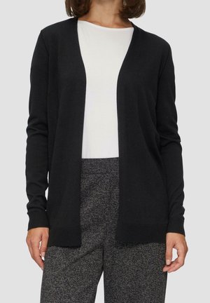 Kvinde iført en sort cardigan med åben front, hvid rundhalset t-shirt og grå teksturerede bukser, stående foran en ensfarvet baggrund.
