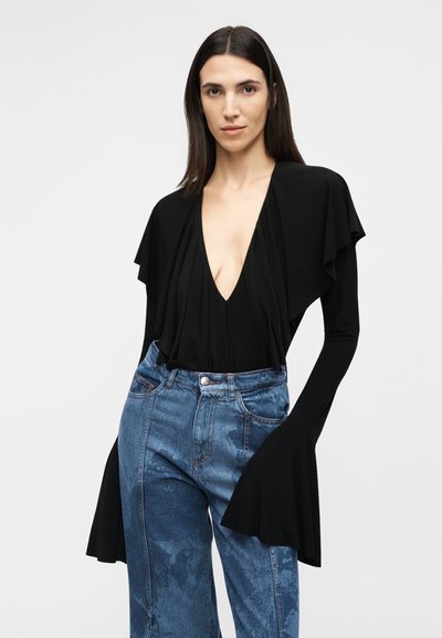 Γυναίκα με ίσια σκούρα μαλλιά φοράει μαύρο τοπ με βαθύ V και φαρδιές μανσέτες, καθώς και ψηλόμεσα μπλε τζιν, πάνω σε ανοιχτό φόντο.