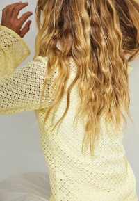 Lys gul, strikket sweater med løs pasform og åbenmønster. Lange, bølgende hår delvist synlige, hånd hævet. Struktureret vævning detail.