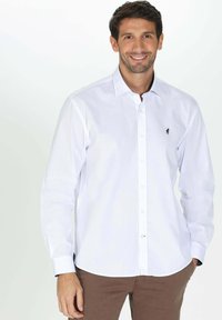 Chemise blanche à manches longues avec un col pointu, arborant un petit logo marine sur la poitrine. Le tissu semble lisse et a une coupe ajustée.